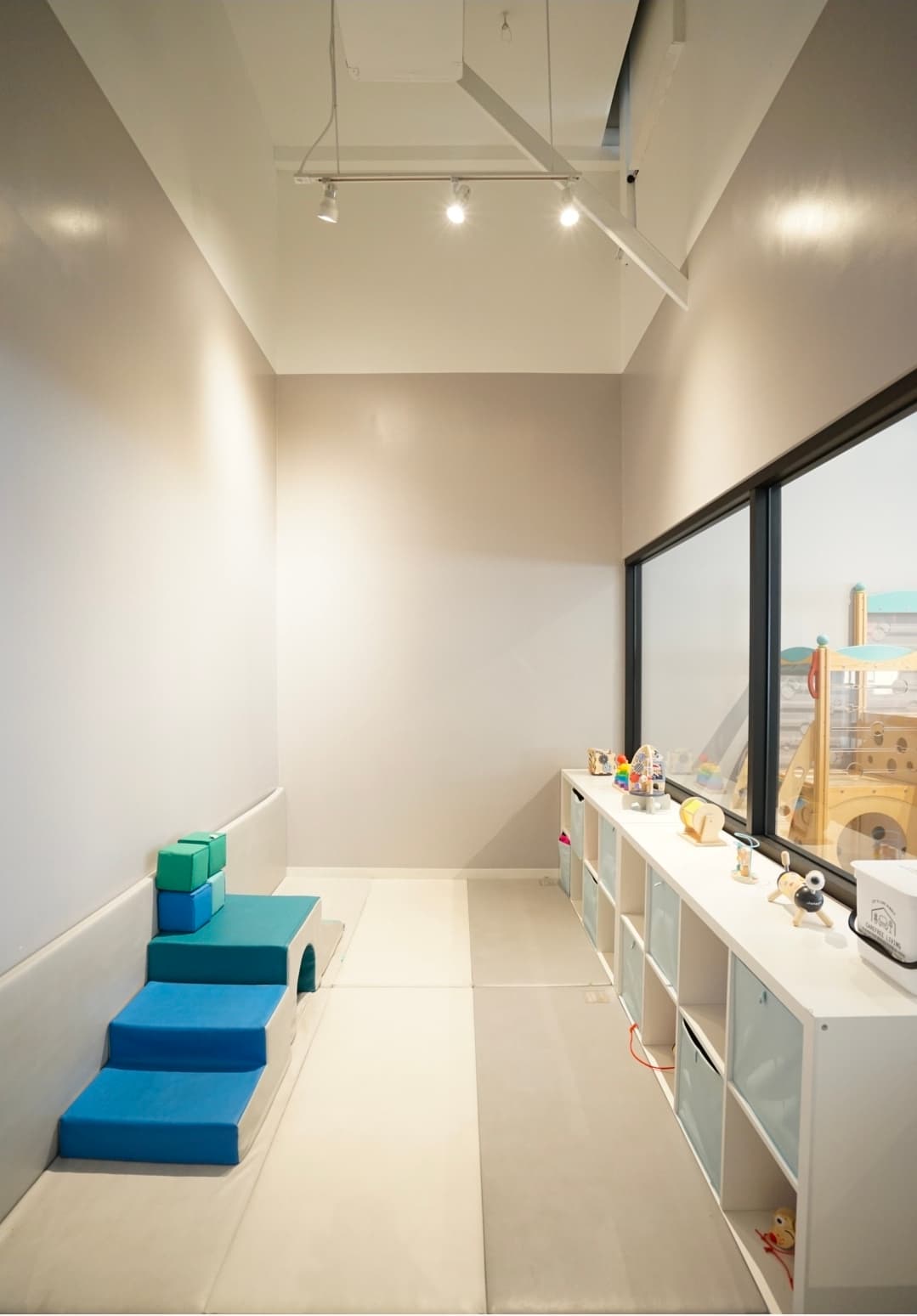 Quiet soft-play room with blue foam steps, floor mats, toy shelves, and a window into the main playground